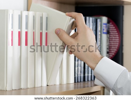 Choosing book. Hand of man in white sleeve cuff shirt taking up grey book out of big range staying in row on book shelf cupboard cabinet home interior