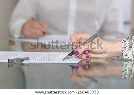 Female hand signing formal paper. Close-up hand on foreground working with paper sheet fountain pen male official dressed body on background glass office desk with reflection woman hand nail make up