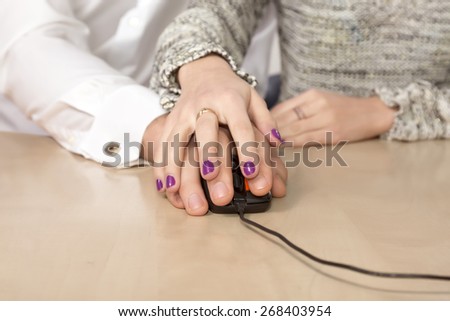 Learning computer skills.\
Female hand over the male hand directing his moves with computer mouse.\
Desk, table, beige, worm, nail make-up on woman fingers. Blurred people body on background.