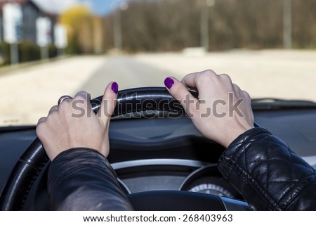 Hands of mature female racer on steering wheel.\
Woman holds the cars steering wheel. Blurred road, forest and blue sky on the background. Skin is not smoothed to emphasize experience of driver