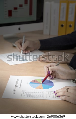 Hands of people working on presentation. Office desk with cropped laptop and large desktop on the foreground, male and female hands keeping pens and pointing on the colored data charts sunbeam
