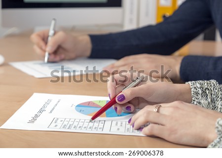 Hands of people working on presentation. Office desk with cropped laptop and large desktop on the foreground, male and female hands keeping pens and pointing on the colored data charts side view