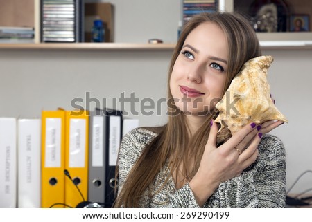 Dreaming lady listening sea shell. Beautiful smiling Caucasian young woman holding large sea shell and listening the sound dreamily looking into far. Office interior, casual dress code