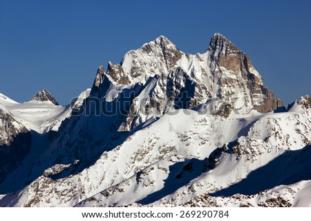 High contrast mountain landscape. Image of part Main Caucasian mountain range with two-headed peak in middle panorama dark flat saw shaped summit on left high contrast blue sky orange red tone rocks