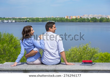 Young couple on parapet looking sidewards. Two young people male and female sitting on parapet embracing in front of urban landscape with river and buildings looking apart together