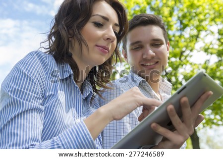 Man and woman with tablet PC on blue sky background. Portrait two people male female young looking on tablet PC talking blue cloudy sky green tree summer background