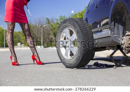 Leggy female changing wheel of car.\
Lady dressed in provocative clubbing pantyhose and bright red shoes high heels mini skirt rolls large wheel to car fixed on jack-screw with some tools