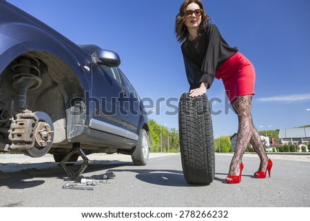 Leggy female changing wheel of car.
Lady dressed in provocative clubbing pantyhose and bright red shoes high heels mini skirt rolls large wheel to car fixed on jack-screw with some tools