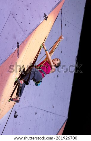 Female extreme climber fixing the rope into belay quick draw. Strong muscular girl in red pants and black top makes a difficult ascent on outdoor climbing wall night spotlight colorful