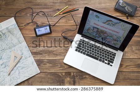 Preparing for trip. Wooden desk at home laptop with map connected to navigation devise sign MAP UPLOAD COMPLETE unfolded detailed topographic map