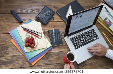 Man working at wooden desk.  Man typing on keyboard drinking coffee at wooden desk with laptop newspaper magazines and plate of strawberry