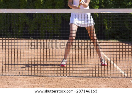 Slim legs of female tennis athlete behind fishnet barrier.\
Young woman stays in ready position close to tennis playground barrier ready to return the ball white dress miniskirt focus on legs