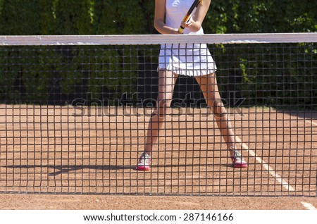 Slim legs of female tennis athlete behind fishnet barrier.\
Young woman stays in ready position close to tennis playground barrier ready to return the ball white dress miniskirt focus on fishnet