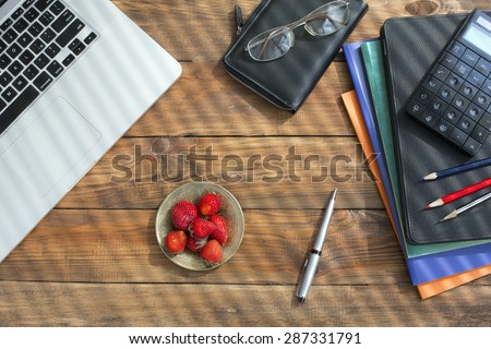 Modern technology and nature concept.
Very natural vintage looking working desk top view grey laptop booklets pen pencils plate fresh strawberry striped by sunbeams coming throw venetian blends