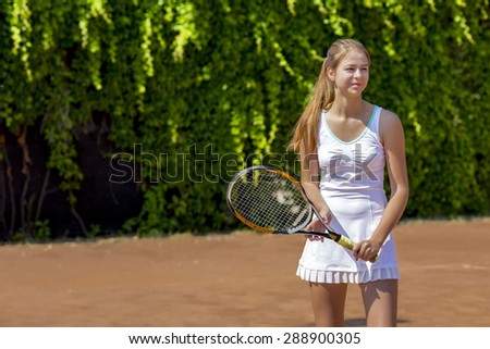 Smiling tennis athlete on play-field.\
Junior female stays on clay tennis court white dress with miniskirt holds racket smiling positive green fence blurred background