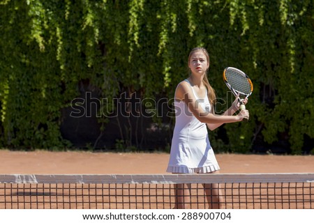 Well prepared to defend. \
Young female tennis athlete swinging racket to meet coming ball white dress miniskirt outdoor play court dark green fence background orange clay ground copy space on right
