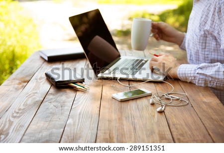 Freelance work.\
Casual dressed man sitting at wooden desk inside garden working on computer pointing with color pen drinking coffee gadgets dropped around on table side view