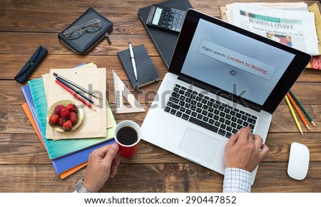 Booking flight tickets on-line concept. Man working on computer booking air flight tickets screen searching best offer from London to Beijing wooden desk stationery books pen pencil color on table