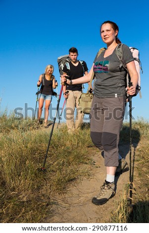 Family of hikers walks on clay path.\
Three people two female one female heavy loaded with backpacks and trekking gear go along path throw wild grassy hill blue sky on background