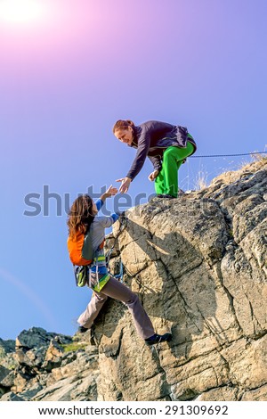 Climbers teamwork.\
Two climbers reaching summit one holding hand of her partner assisting to make last step to top young female athletes natural stone rock blue sky and shining sun on background