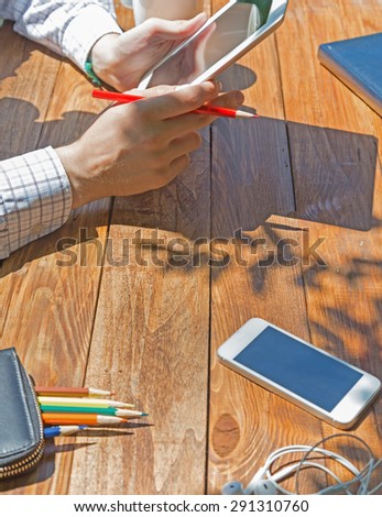 Wooden desk and man browsing gadget. Brown natural wood table under shining sunbeams outdoor electronic devises pencils notepads  around male hands holding tablet PC shadows dropped by tree leaves