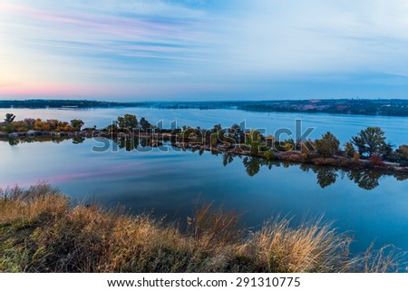 Tenderly colored sunset landscape at East European waterfront. Majestic view of large river with unusual shape island in middle small town buildings on remote waterfront soft color rose red dusk sky