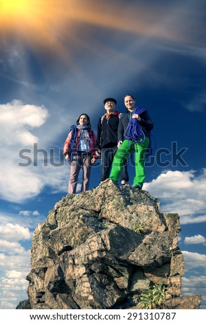 Group of climbers on sharp summit. Family of three people male grandfather and two female daughters having climbing gear stays on rocky cliff blue sky and shining sun background