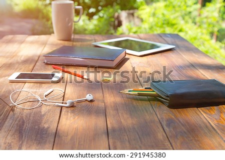 Modern lifestyle background.\
Country rough wooden desk and lifestyle hobby items dropped around electronic devises pencils coffee mug notepad green flora and back light sunbeams behind