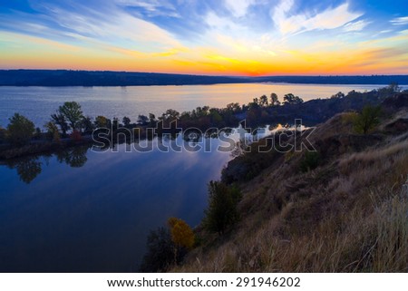 Gorgeous dawn on blue lake.\
Luminous sunrise blooming over wild waterfront landscape with fall grass flower hill stone rocks and narrow island with colored trees in the middle of water surface