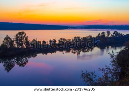 Amazing orange dawn on blue lake.\
Luminous sunrise blooming over wild waterfront landscape with fall grass flower hill stone rocks and narrow island with colored trees in the middle of water surface