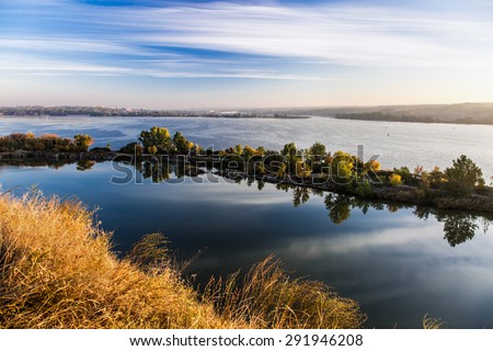 Suburban East European waterfront landscape.\
Wide river with unusual shaped narrow island in middle of water surface blooming yellow grass flowers color tree cloud sky background