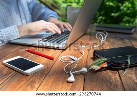 Man arms typing on keyboard at natural hardwood desk with green flora background many electronic gadgets dropped around tablet PC smart phone color pencils headphones  coffee mug sunlight day outdoor