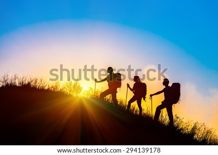 Silhouettes of three people walking with backpacks and other hiking gear up toward top of wild grass mountain mother father daughter bright luminous sunrise sky background