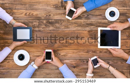 Group of Business People on Informal Meeting at natural wood Cafe Desk using variety of electronic gadgets connecting to internet and accessing data wireless two coffee mugs at corners