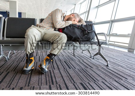 Man stuck at airport.\
Image of bearded informal style dressed person sleeping on his travel backpack inside airport waiting lounge sitting in black chair with heavy alpine boots on his legs