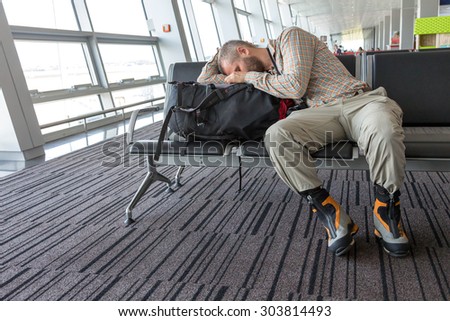 Man stuck at airport.\
Image of bearded casual style dressed person sleeping on his travel backpack inside airport waiting lounge sitting in black chair with heavy alpine boots on his legs