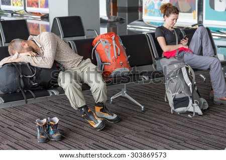 Passengers waiting for the air flight at airport terminal.\
Man and woman sitting in chairs line sleeping browsing smart phone internet casual dress code heavy boots backpacks luggage building interior