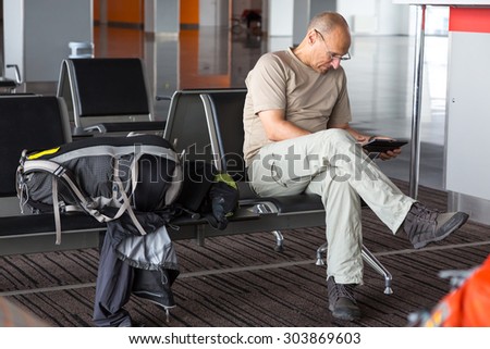 Aged passenger waiting for boarding.\
Mature man using tablet PC sitting inside airport terminal building chair line interior day informal simple dress code large cabin luggage sport backpack