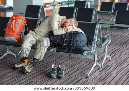 Sleeping man inside transport terminal.\
Man couching on his luggage waiting for train departure sport smart casual relaxed dress code soft shirt and pants heavy winter boots interior background