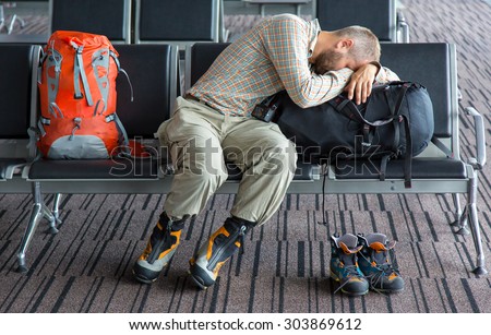Difficult journey.\
Man sleeping on his luggage lean onto large backpack heavy winter boots and smaller pair aside airport terminal building interior