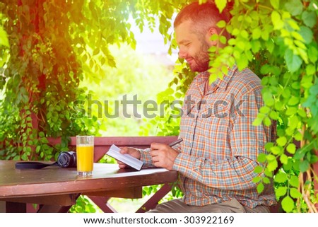 Travel diary.
Bearded man sitting at vintage design wooden arbor and writing notes into his notepad having glass of orange juice and photo camera on table surrounded green flora sun light background