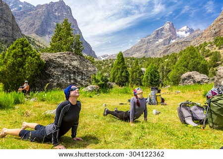 Young women doing morning fitness in mountain landscape.\
Two female athletes on green grass lawn doing yoga sport exercises outdoor day sunlight blue sky and mountains on background