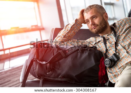 Traveler looking into far.\
Portrait of bearded man inside airport terminal pensive casual shirt and travel pants sitting next to his backpack luggage large bright window on background