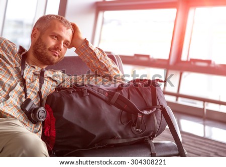 Smiling man at airport. \
Positive bearded passenger sitting airport lounge waiting for his flight casual dress shirt pants large backpack luggage photo camera sun light coming throw windows background