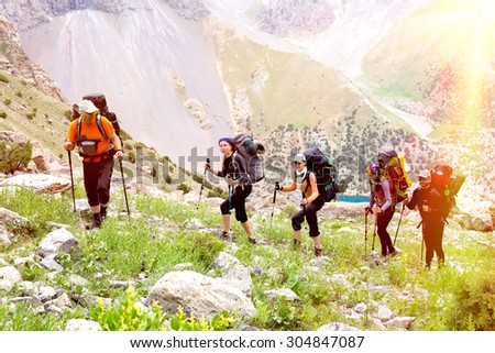 Group of people walking on trail.\
Men and women going up with backpack luggage and hiking gear on bright mountain landscape background with sun rising and high peaks behind