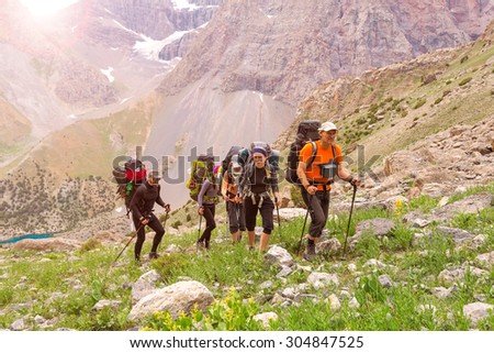 Group of people walking on trail.\
Men and women going up with backpack luggage and hiking gear on bright mountain landscape background with sun rising and high peaks behind