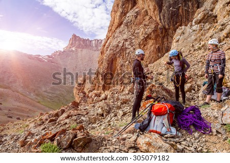 Mountain climbers preparing for ascent Three people male female placing gear packing backpacks staying on rocky terrain at beginning of Climbing Route on Mountains blue Sky and Sun Rising Background