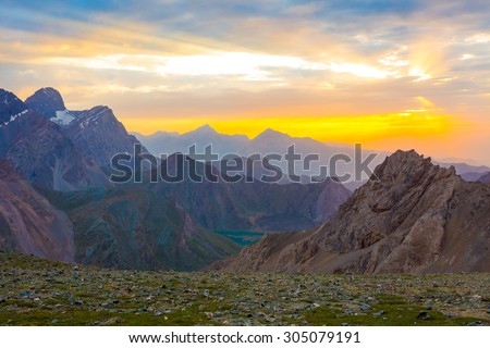 Eye catching highland dusk. \
Beautiful eye catching mountain scenery with rising sun sunbeams passing throw clouds green grass and flowers on foreground brown and red peaks and ridges on background