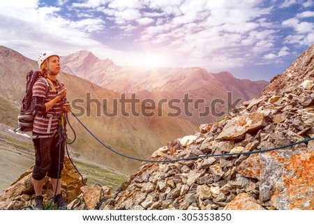 Mountain climber working with rope. Young woman belays her climbing partner with rope keeping it with both hands have protection gear helmet safety harness mountain landscape and rising sun background