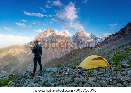 Explorer talking on satellite phone.\
Silhouette of man in wild mountain landscape walk along yellow camping tent holding radio transmitter connection with team blue sky sunny evening